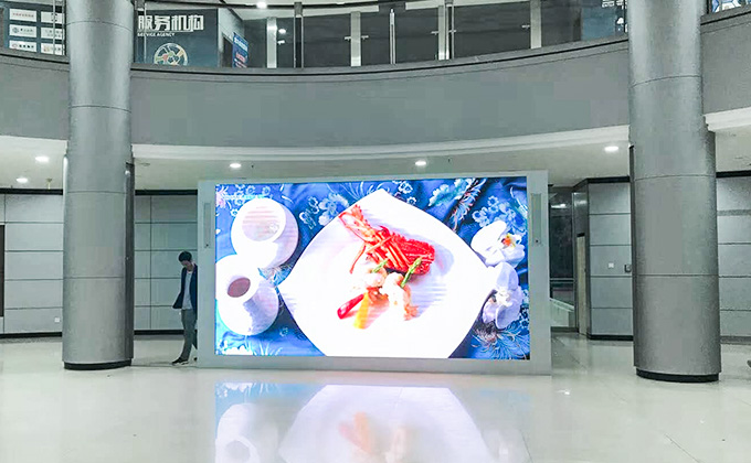 In a lobby, a large LED display (led screen) shows a vibrant food dish. With a person walking by, it highlights the led screen\'s role in indoor visual attraction and information display.