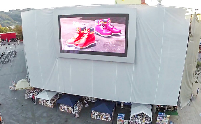 An outdoor LED billboard (led display screen) shows colorful boots ads on a large building. With stalls below, it highlights the led screen\'s role in outdoor advertising, capturing public attention.